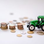 Trator verde em miniatura ao lado de pilhas de moedas, em uma composição ilustrativa sobre financiamento agrícola.