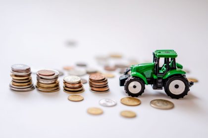 Trator verde em miniatura ao lado de pilhas de moedas, em uma composição ilustrativa sobre financiamento agrícola.