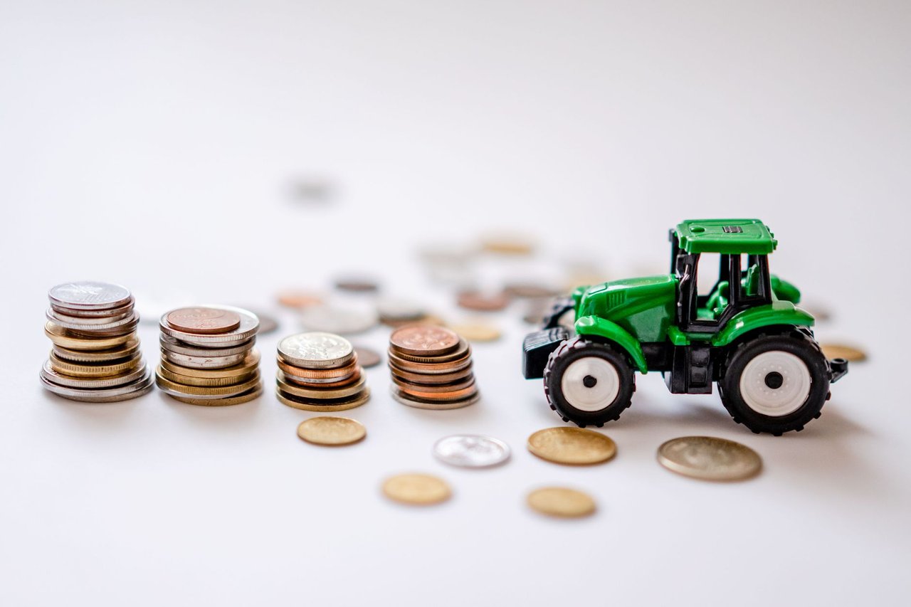 Trator verde em miniatura ao lado de pilhas de moedas, em uma composição ilustrativa sobre financiamento agrícola.