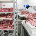 Trabalhadores manipulam cortes de carne em bandejas dentro de uma unidade industrial de processamento de alimentos.
