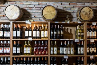 Adega rústica com garrafas de vinho alinhadas diante de uma parede de tijolos e barris de madeira.